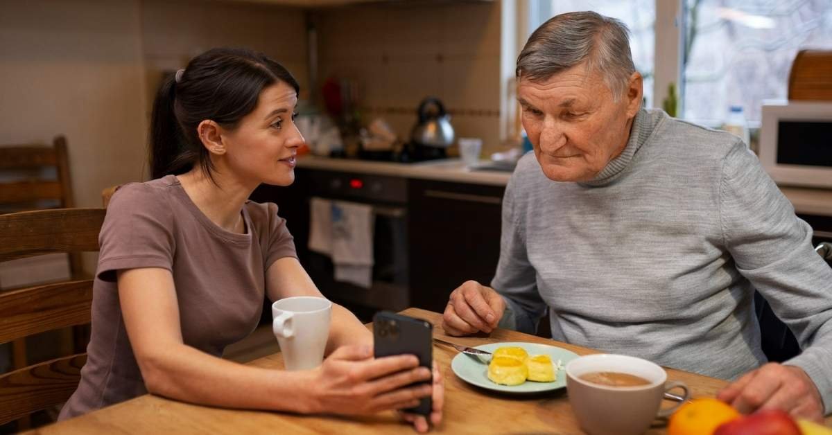 Adult daughter showing her elderly father something on a smartphone while having breakfast at home - caring for parents at home with love and family support.