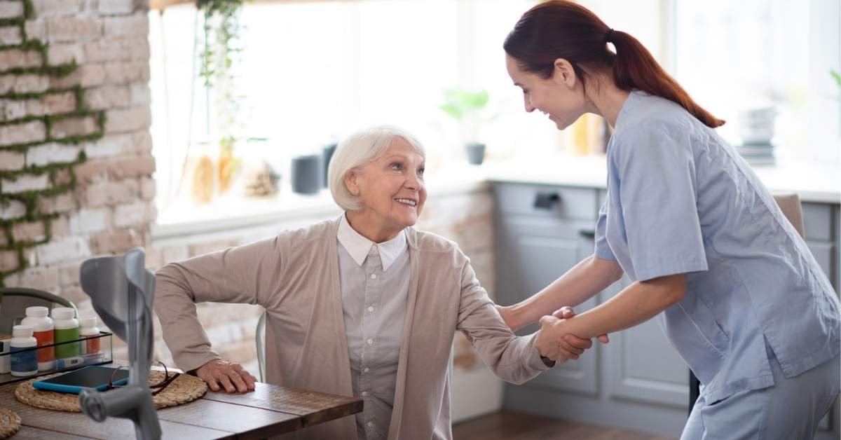 Caregiver helping an elderly woman stand up at home, illustrating home care support for seniors recovering after hospitalization.